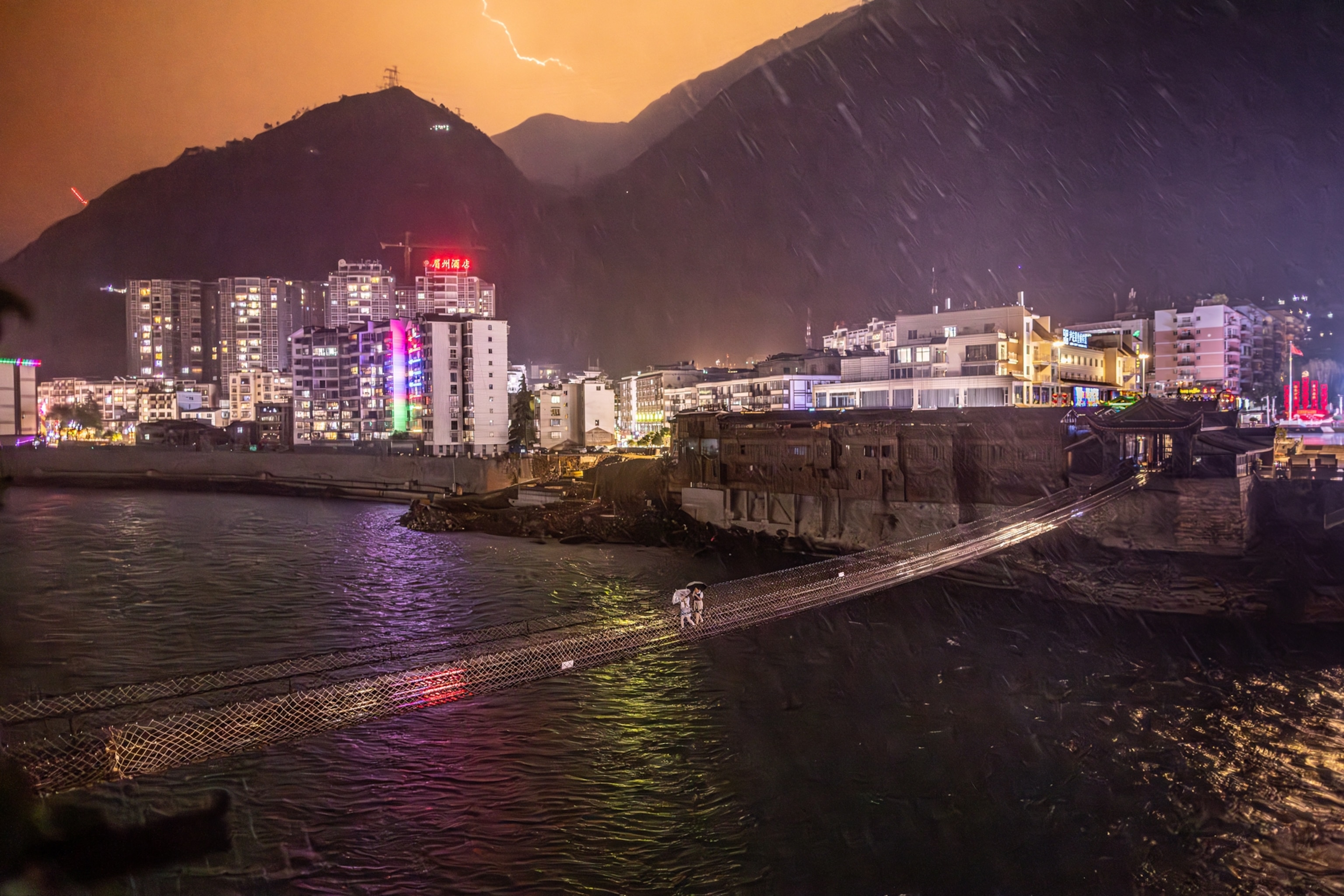 pedestrians walk across a small foot bridge over water in a rainy night