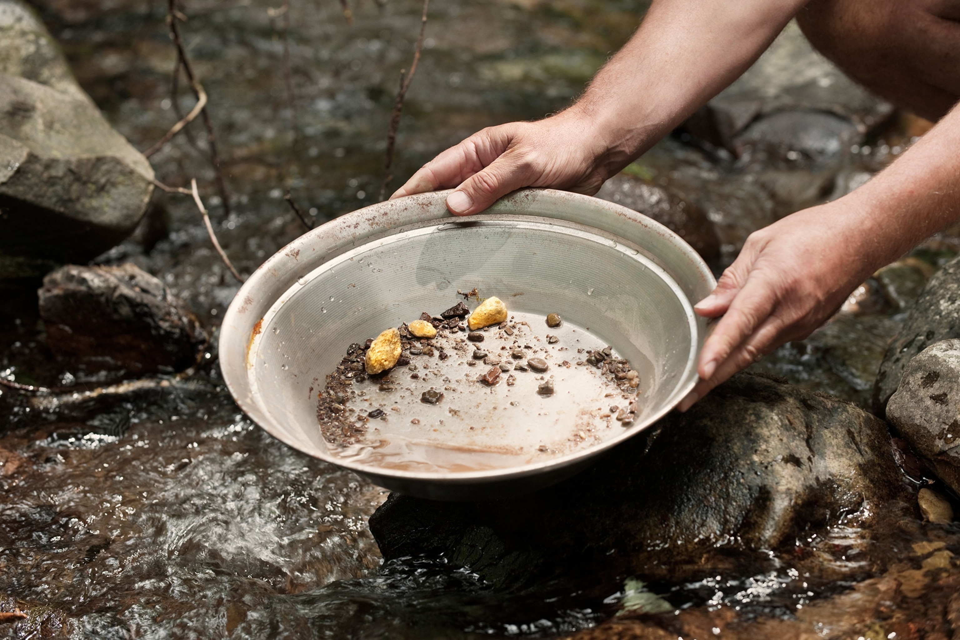 A silver plate filled with rocks and a few pieces of gold nuggets is held over a creek with flowing water.