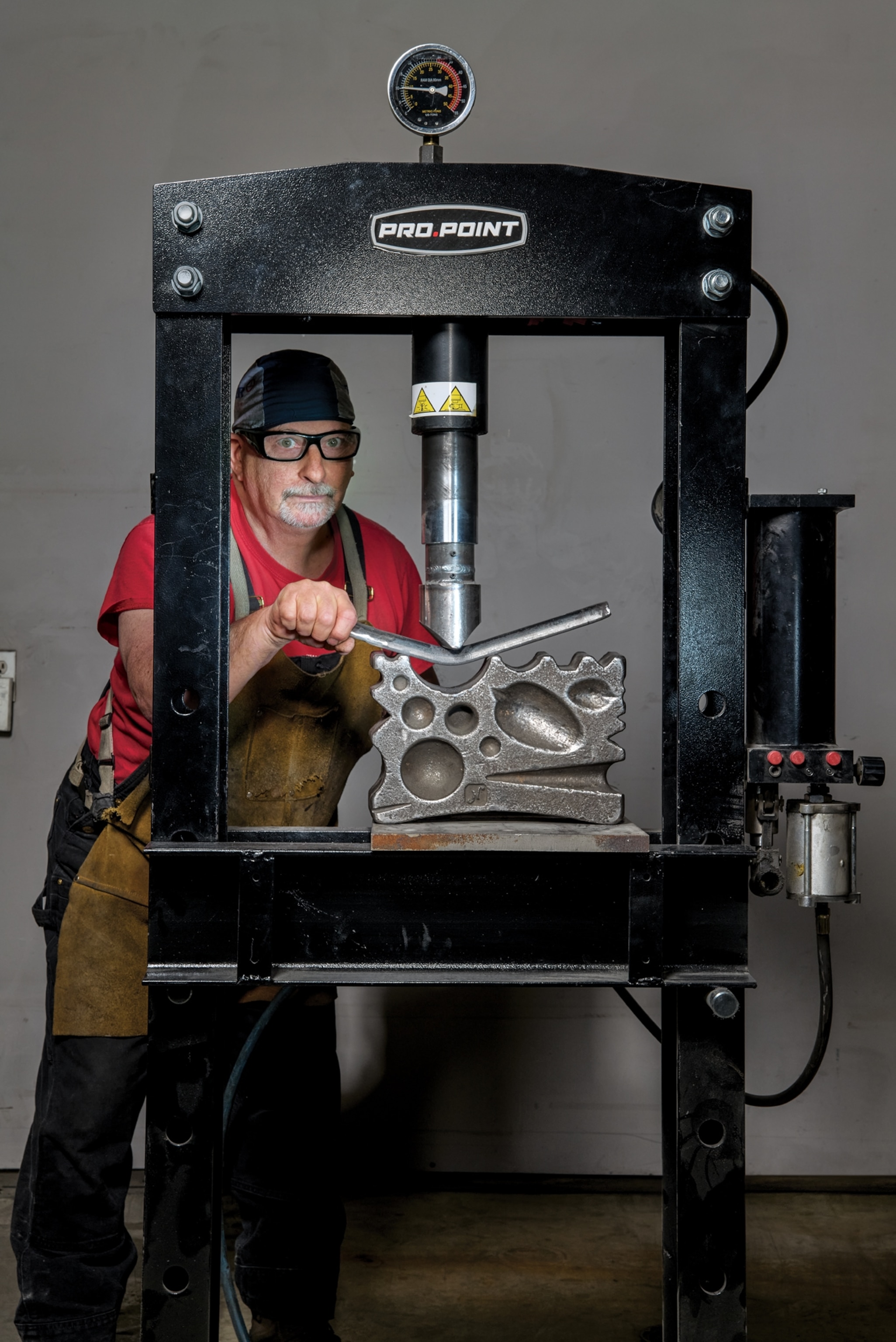 A man wearing protective eyewear, a red t-shirt, jeans, and a work apron poses with a Pro Point swage block machine.