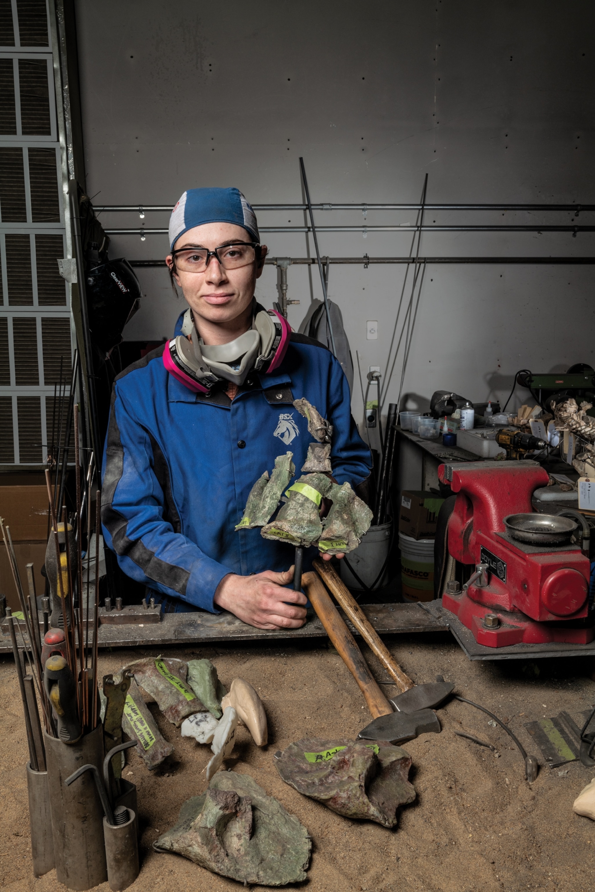 A person wearing a blue jumpsuit and respirator mask around their neck poses holding dinosaur fossils which are fitted together by metal armature.